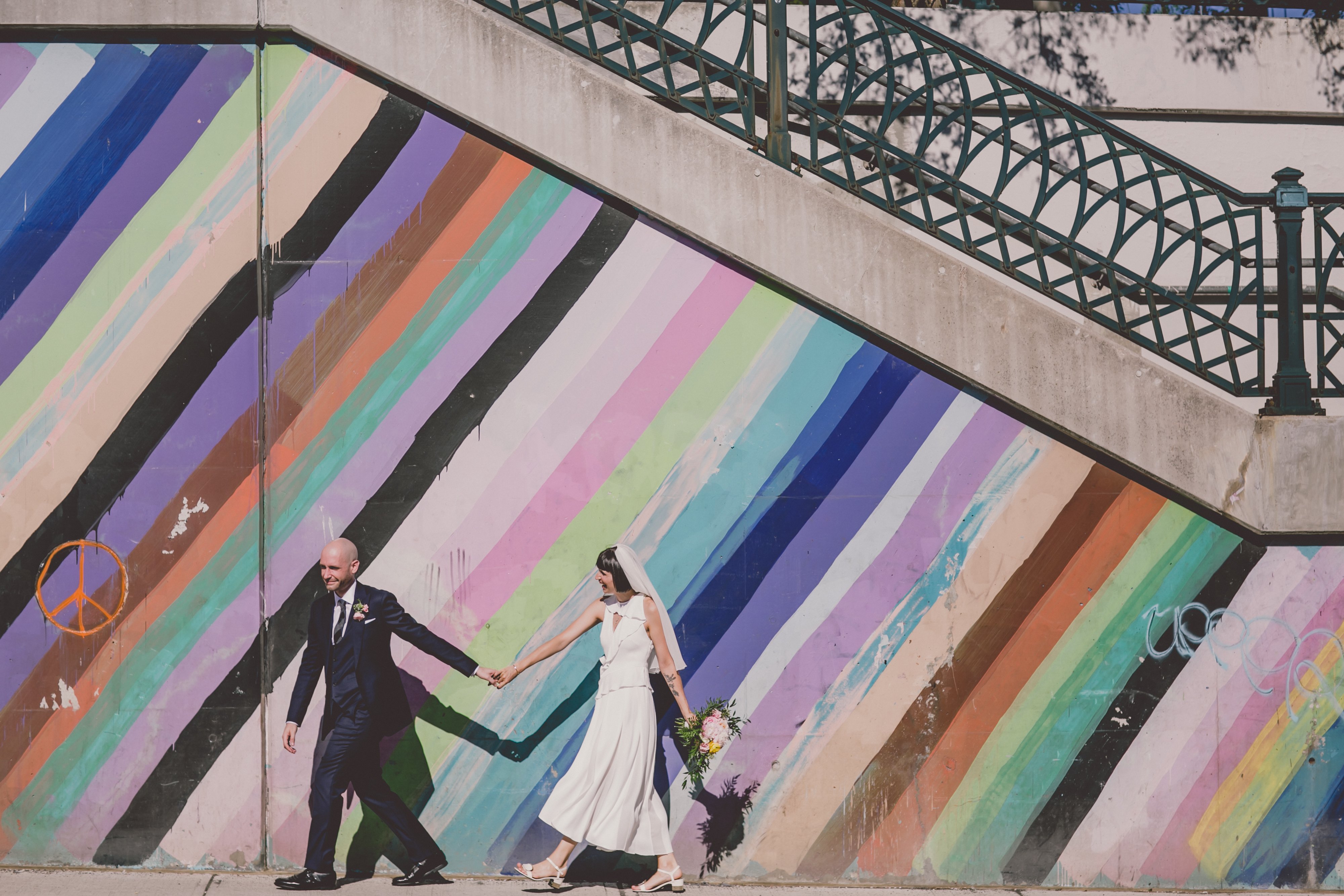 groom leads bride down sidewalk in front of graffiti wall in Roncesvalles.