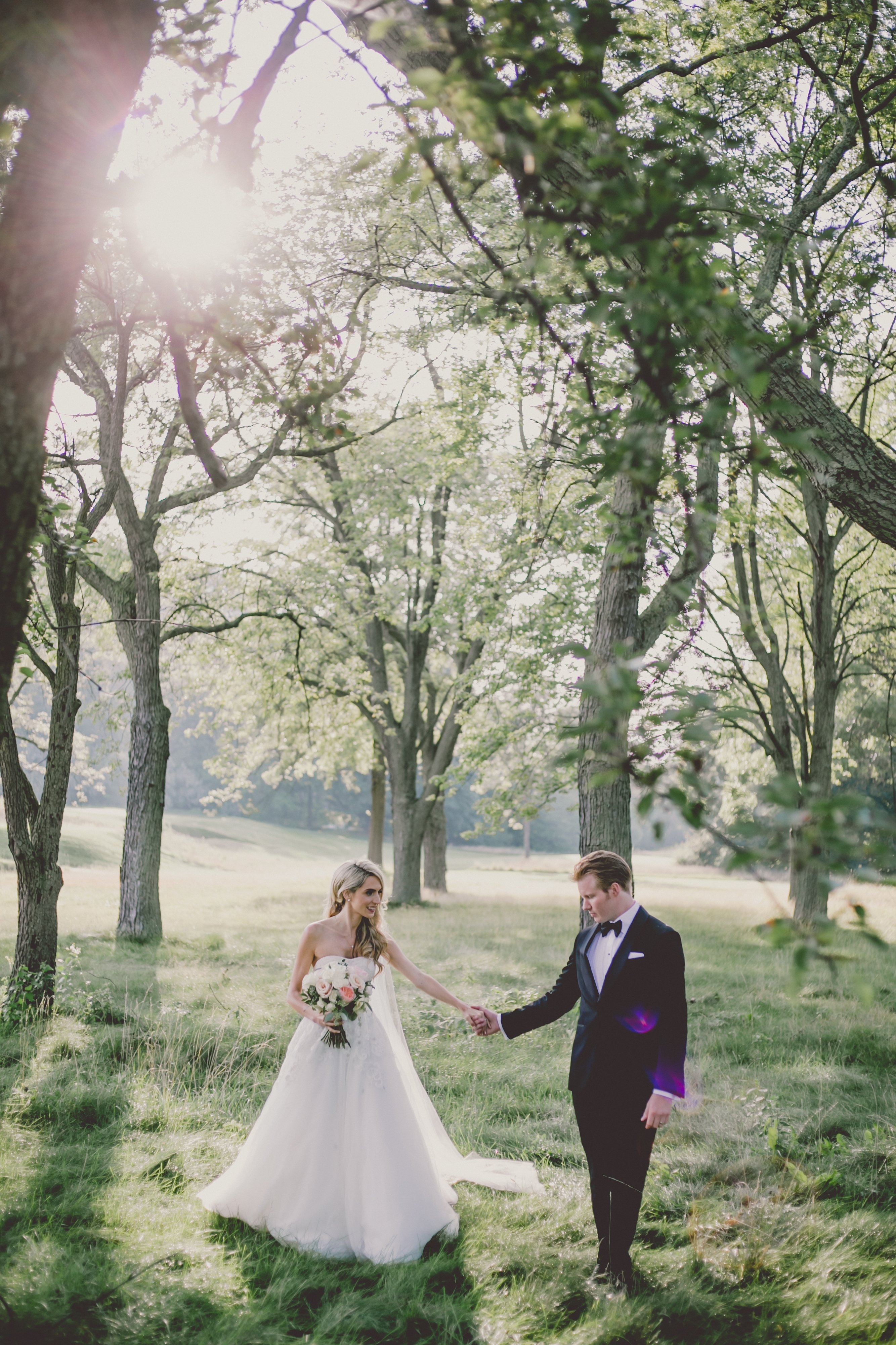 bride and groom walk through grass on summers day with big trees around them