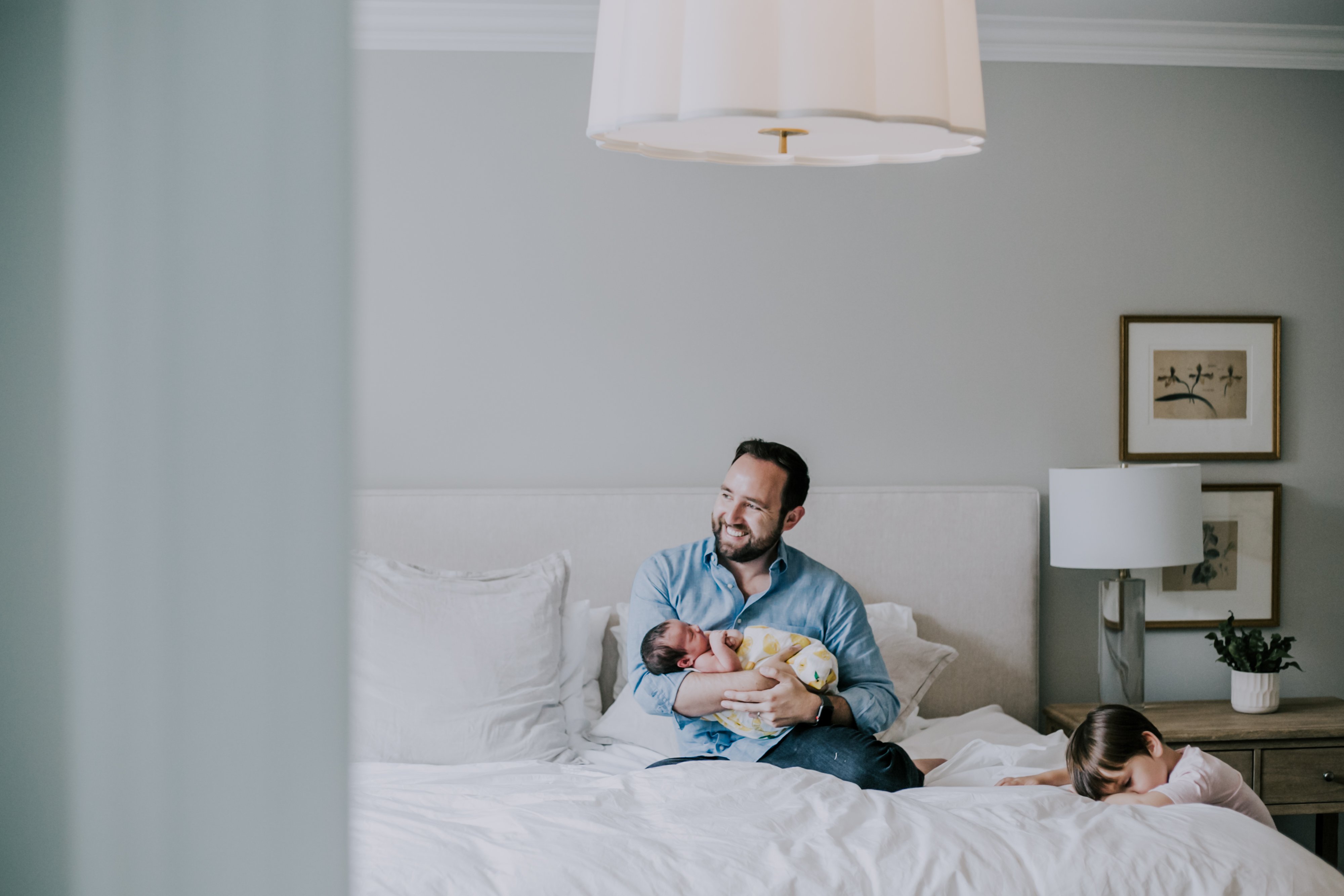 father smiles out window while holding baby sitting on bed