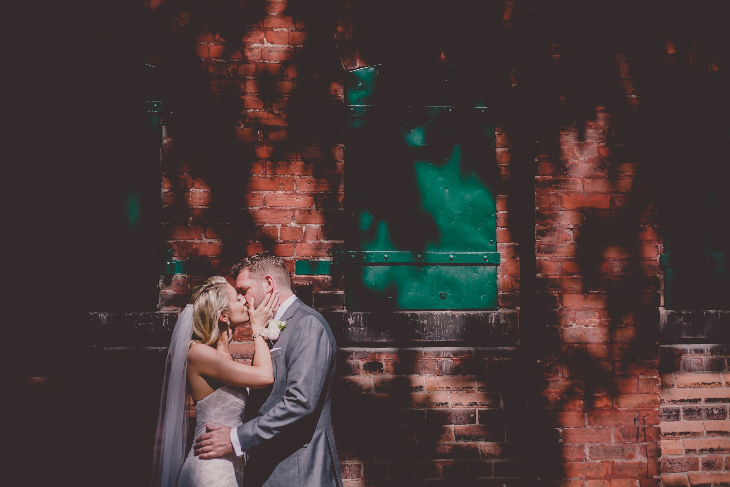 bride kisses groom in front of brick wall while standing in the sun at the Distillery district
