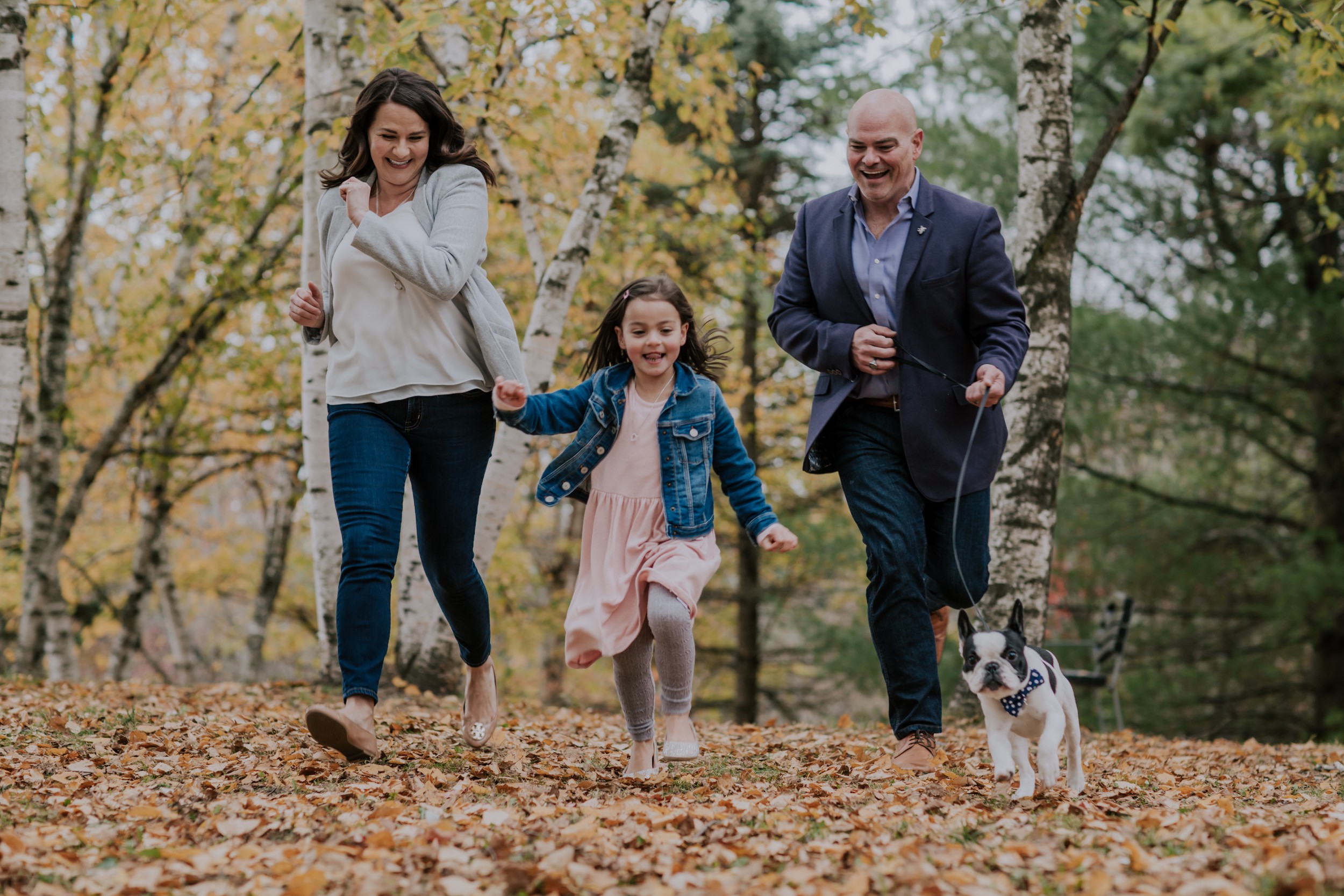 mom, dad, daughter and dog run towards camera lens on fall day