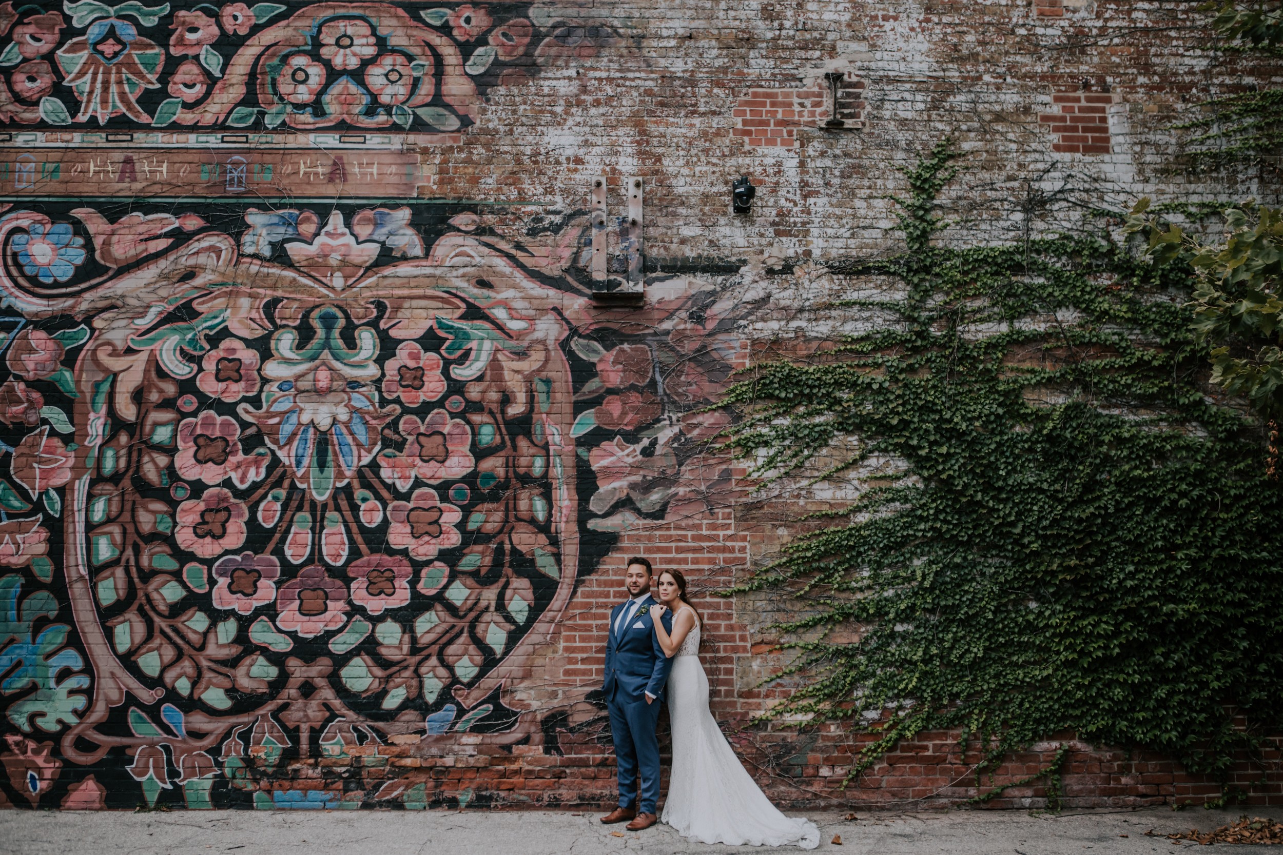 bride hugging groom from behind while standing against a graffiti brick wall