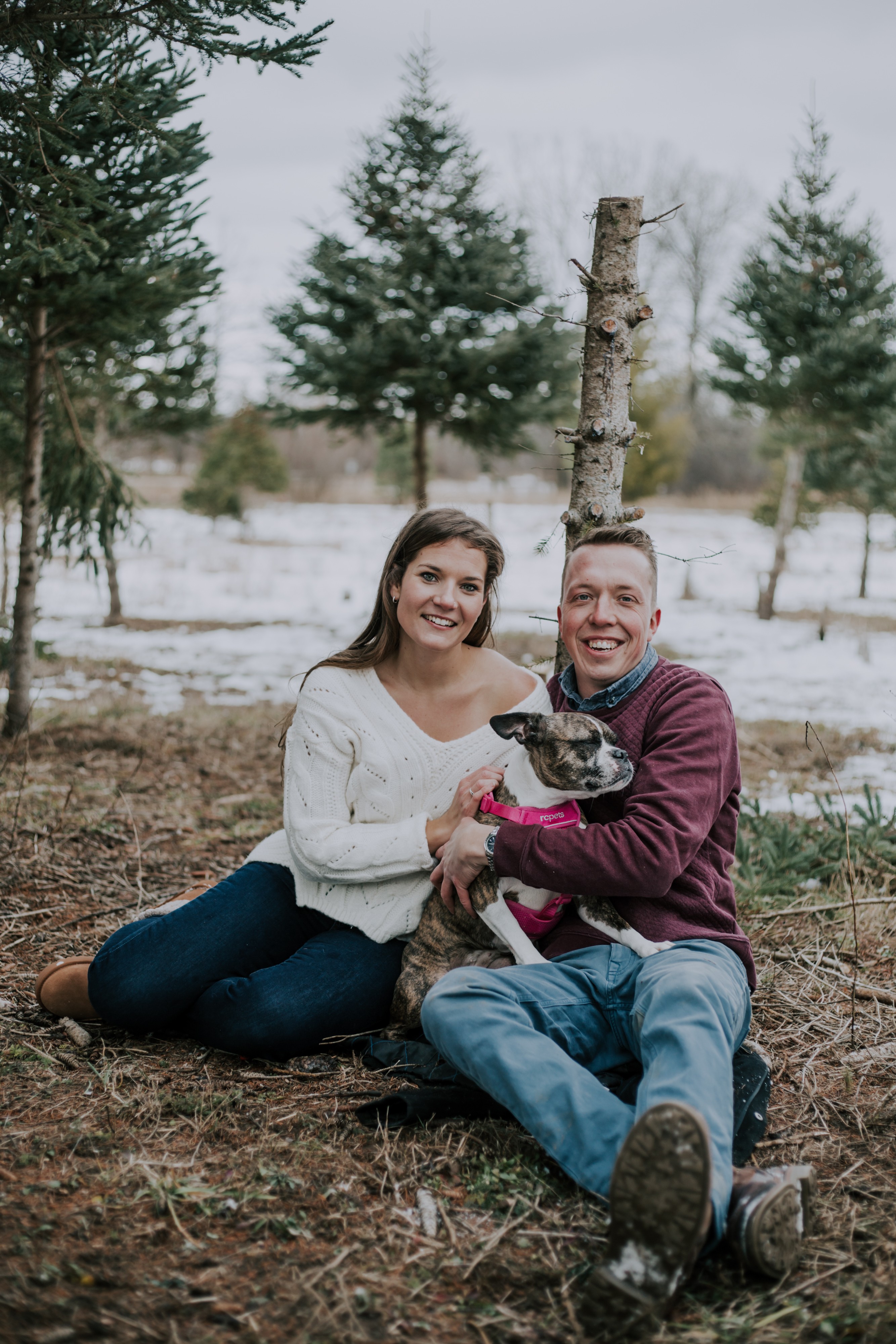 a dog sits on a couple's lap with pine trees and snow behind