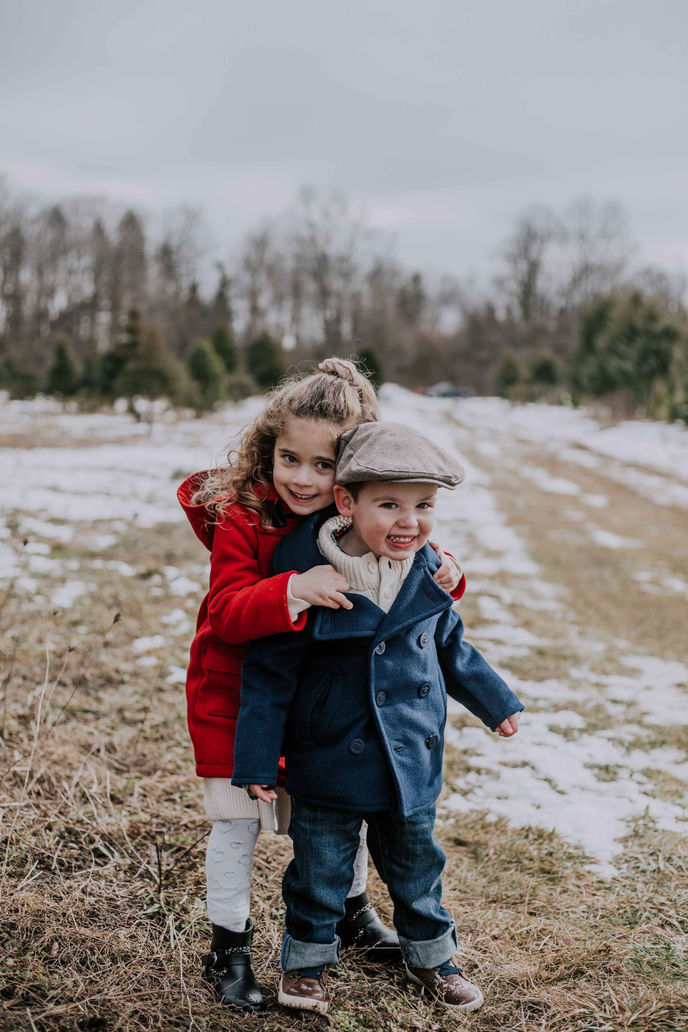 a young girl hugs her toddler brother from behind in a grassy and snowy field