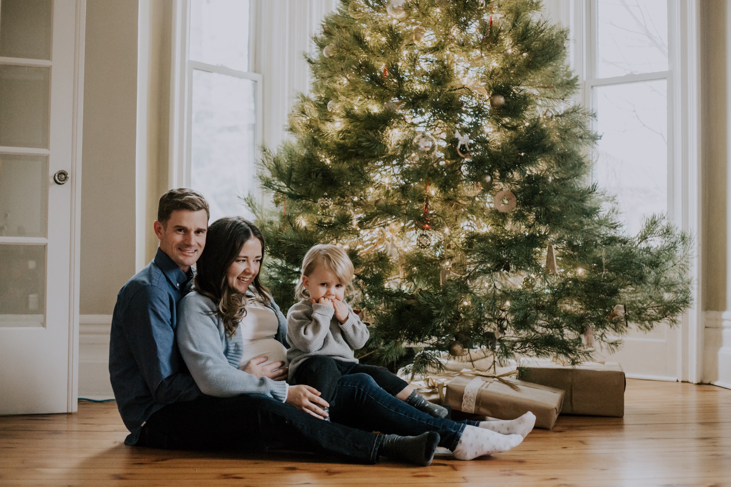 a man sits on floor with wife inbetween his legs and daughter inbetween her legs, all while infront of Christmas tree