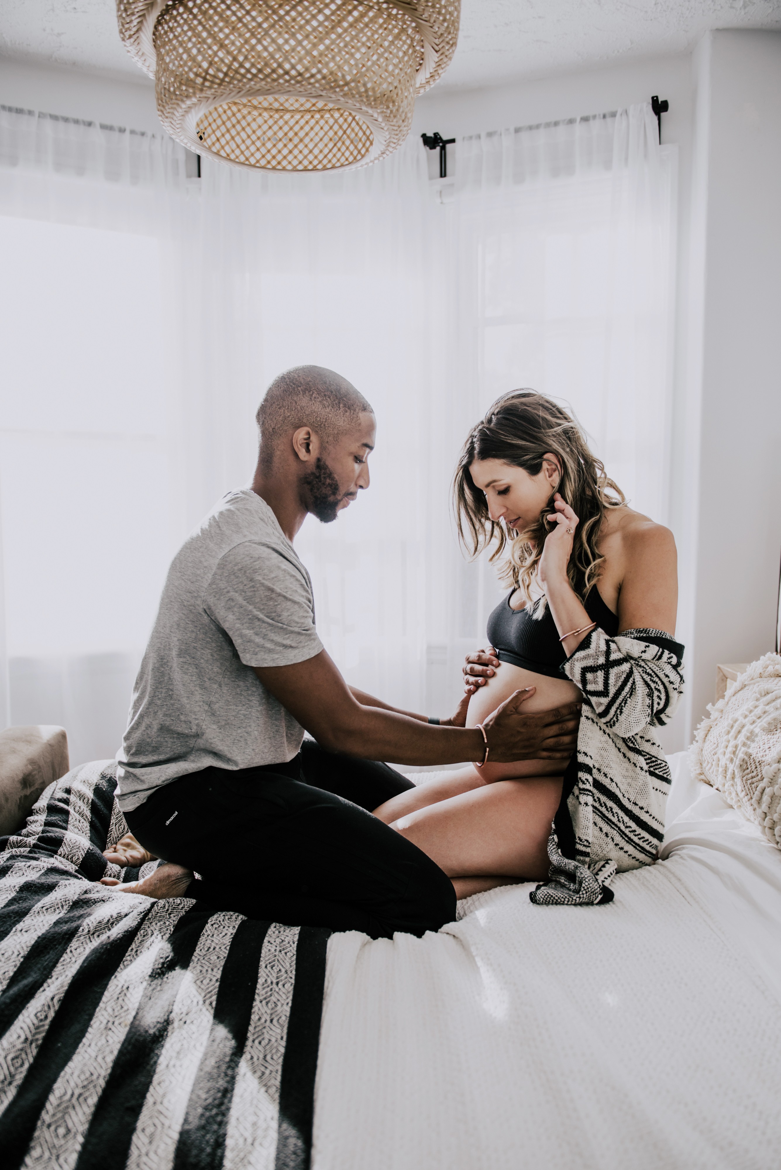 black man touches his white wife's pregnant belly while they kneel on white duvet