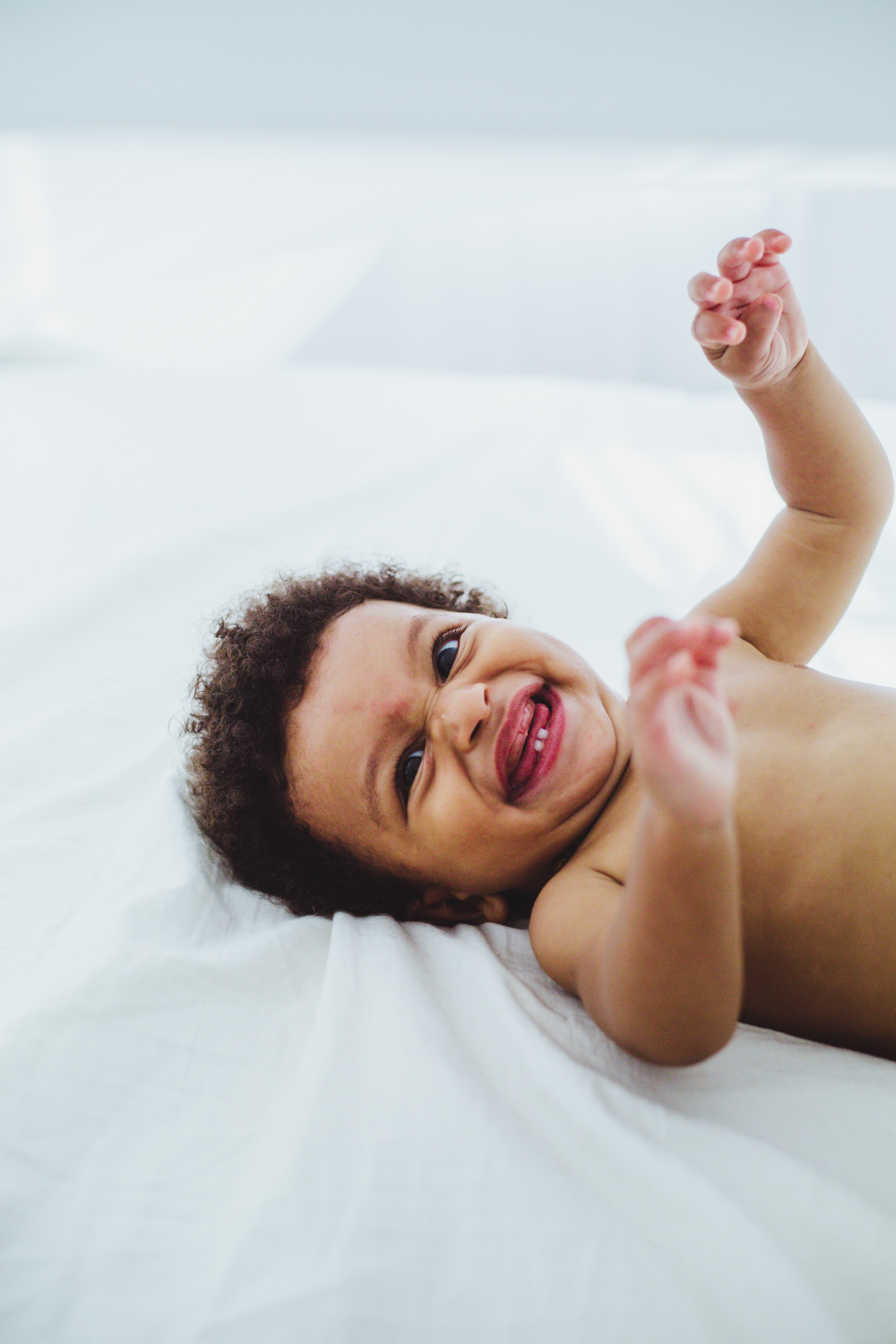 baby boy in diaper laughs towards camera while playing with bed sheets