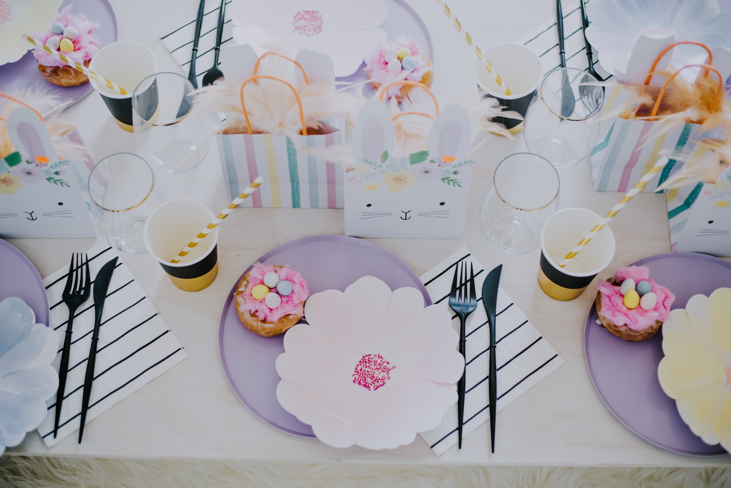 a table top filled with lavender plates, flower plates. black cutlery and bunny gift bags filled with feathers
